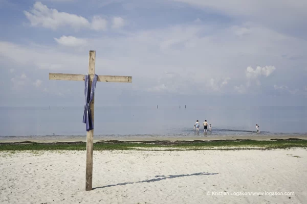 Wooden cross on a beach and in the distans is a family playing in the ocean
